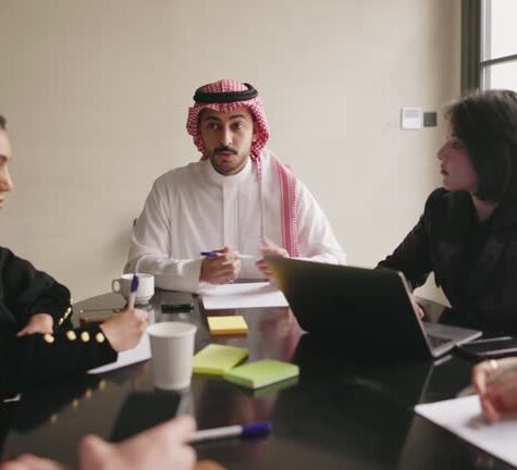 istockphoto-1393567488-640x640 Young businessman in traditional Saudi attire sitting at head of conference table and talking with male and female colleagues about plans in slow motion.